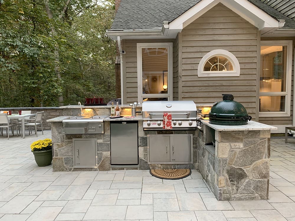 A spacious outdoor kitchen with a stone countertop featuring a built-in stainless steel grill, green egg smoker, sink, and mini refrigerator. The kitchen is set on a stone patio with a table and chairs in the background, surrounded by trees.